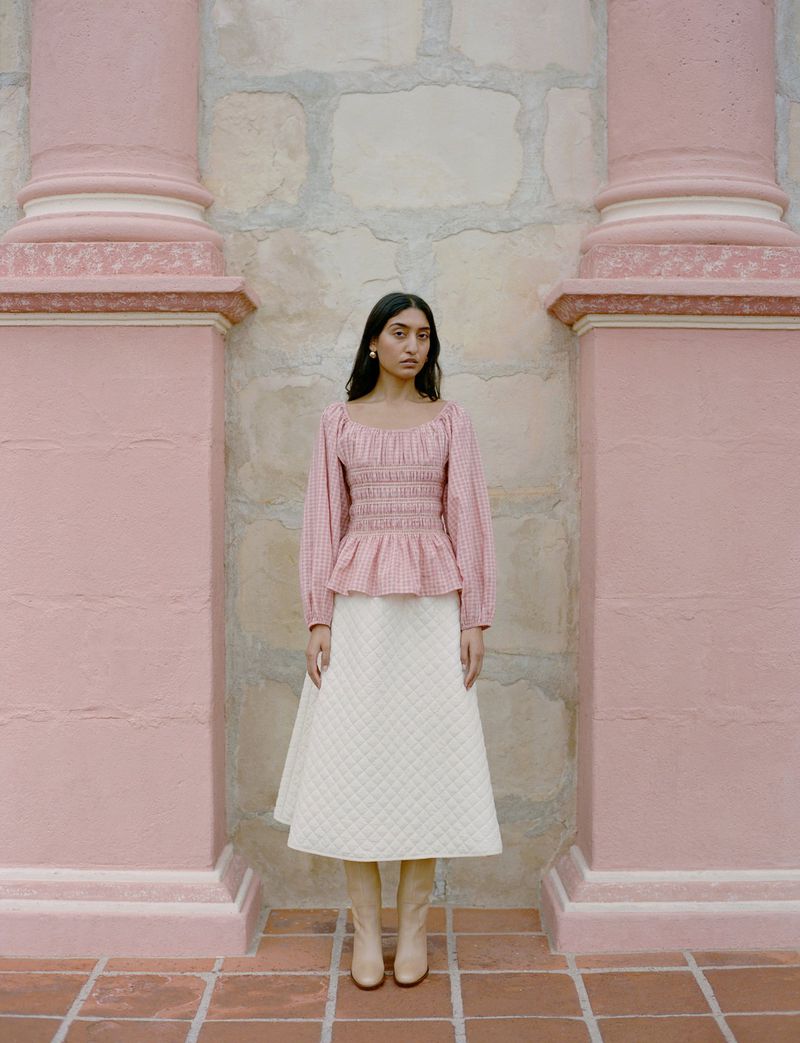 Elegant woman in a pink blouse and white skirt poses against an ornate pink wall. The floor is brick.