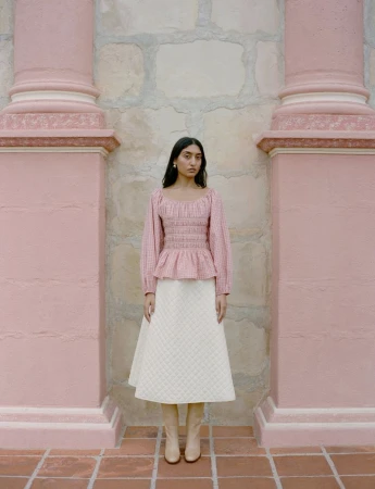 Elegant woman in a pink blouse and white skirt poses against an ornate pink wall. The floor is brick.