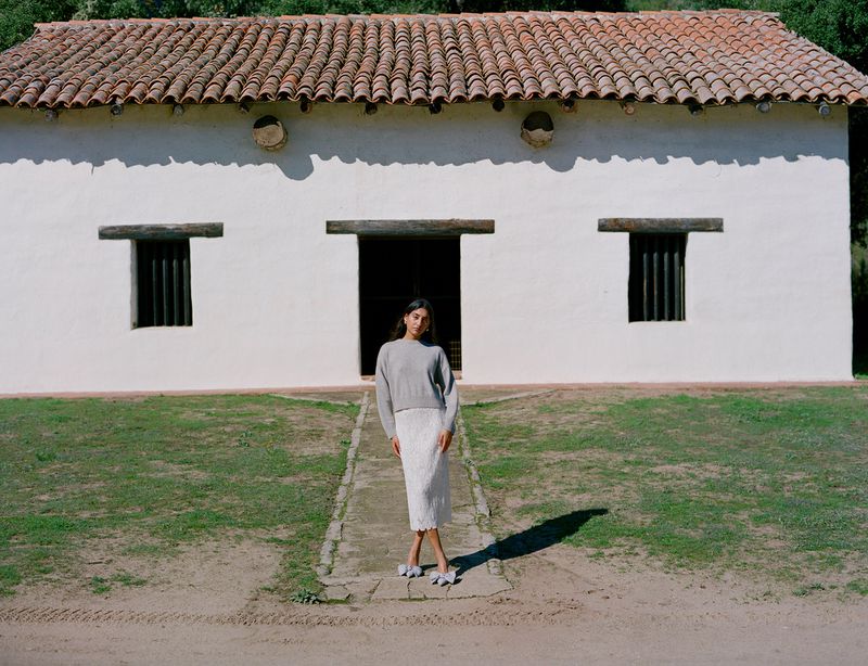 An adult female poses on a path in front of a white building with a red tile roof, wearing a grey sweater and white skirt. Outdoor setting.