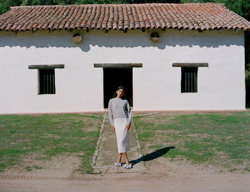 An adult female poses on a path in front of a white building with a red tile roof, wearing a grey sweater and white skirt. Outdoor setting.