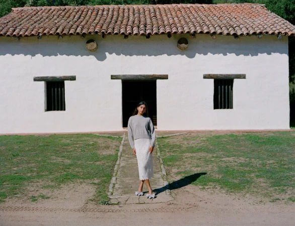 An adult female poses on a path in front of a white building with a red tile roof, wearing a grey sweater and white skirt. Outdoor setting.