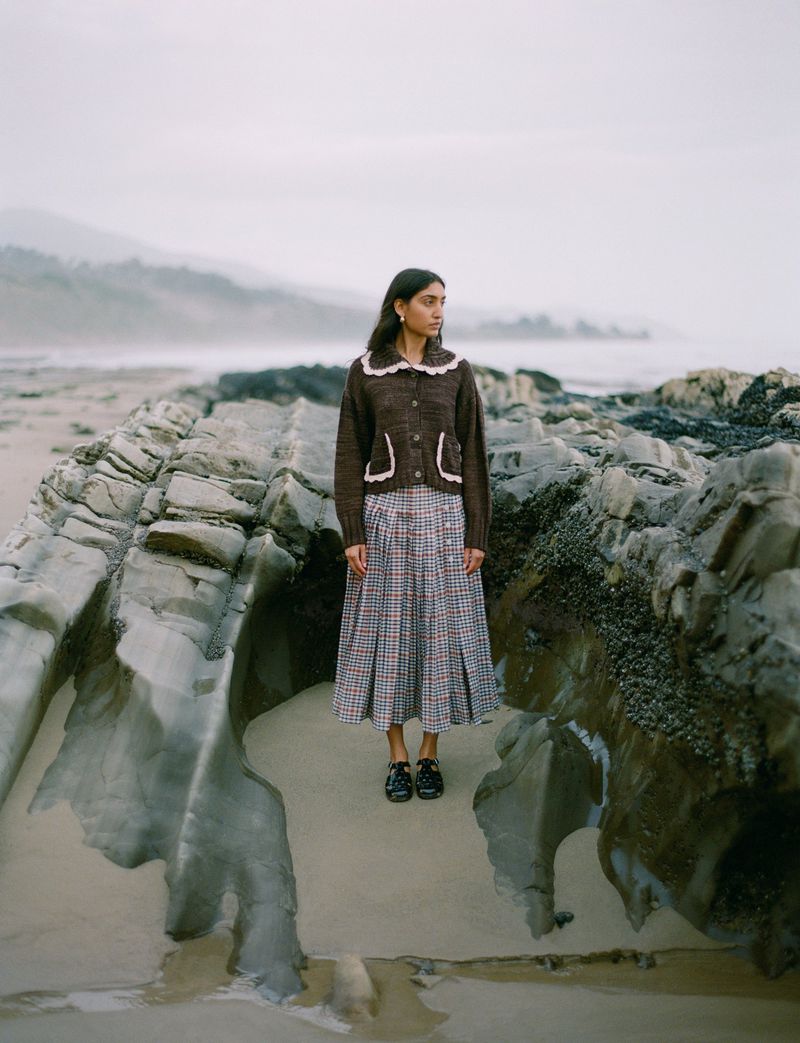 Model standing on rocky coastal shore wearing a brown cardigan and patterned skirt for Loeffler Randall FW24