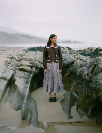 Model standing on rocky coastal shore wearing a brown cardigan and patterned skirt for Loeffler Randall FW24