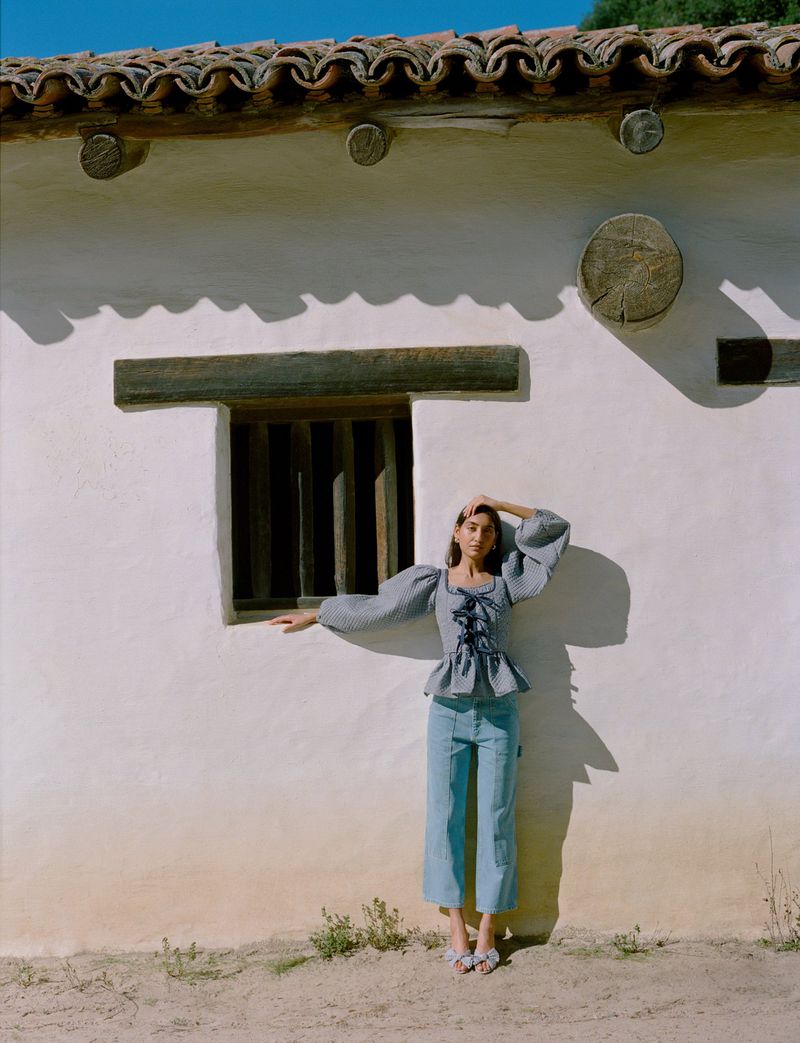 A woman in a blue and white gingham blouse and blue pants poses near a white stucco building with a red tile roof on a sunny day.