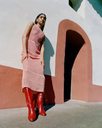 A woman models a pink knit dress and red leather boots outdoors against a white wall with a terracotta archway.