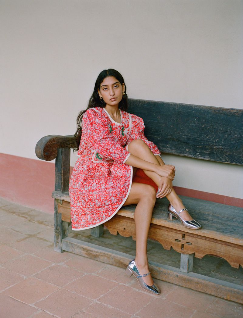 Fashion portrait of a brunette woman in a red floral dress sitting on a bench outside, wearing silver shoes with a neutral wall backdrop.