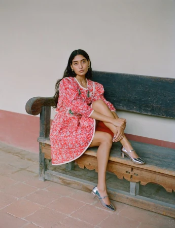 Fashion portrait of a brunette woman in a red floral dress sitting on a bench outside, wearing silver shoes with a neutral wall backdrop.