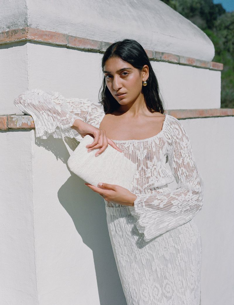 Model in a white lace dress leaning against a stucco wall while holding a matching clutch bag