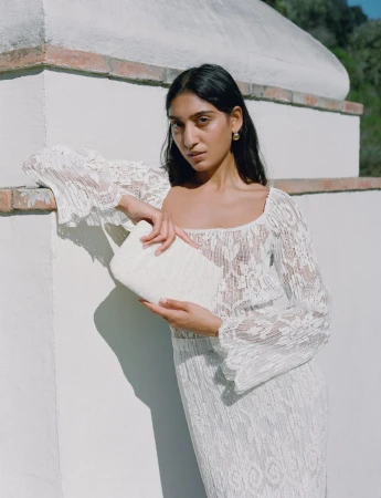Model in a white lace dress leaning against a stucco wall while holding a matching clutch bag