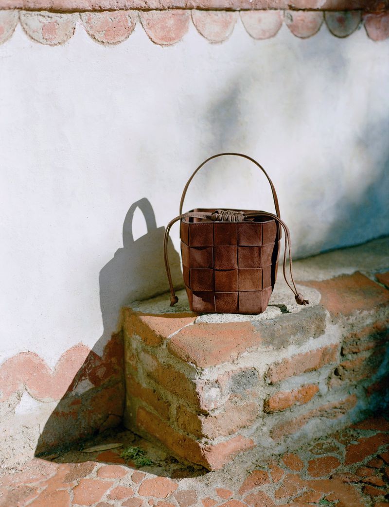 Brown woven bucket bag with drawstring closure and handle, resting on a tiered brick surface outdoors against a stucco wall.