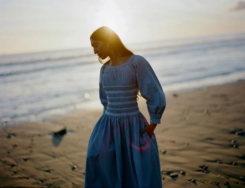 Woman in blue dress standing on beach at sunset with ocean background, looking down. Fashion shoot in natural light.