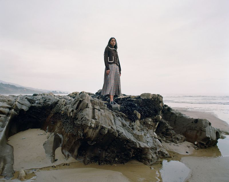 A lone woman stands on weathered rocks by the ocean, wearing a dark jacket and long skirt. Foggy sky and peaceful scene.
