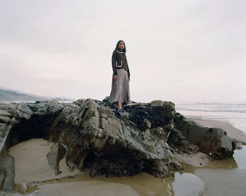 A lone woman stands on weathered rocks by the ocean, wearing a dark jacket and long skirt. Foggy sky and peaceful scene.