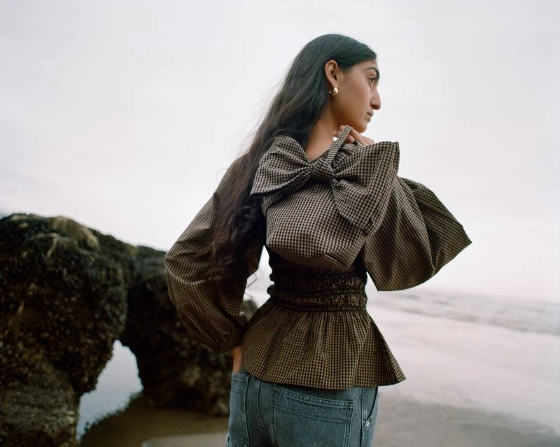 Woman in a brown and white checkered top with bow detail and blue jeans poses on the beach next to rocks on an overcast day.