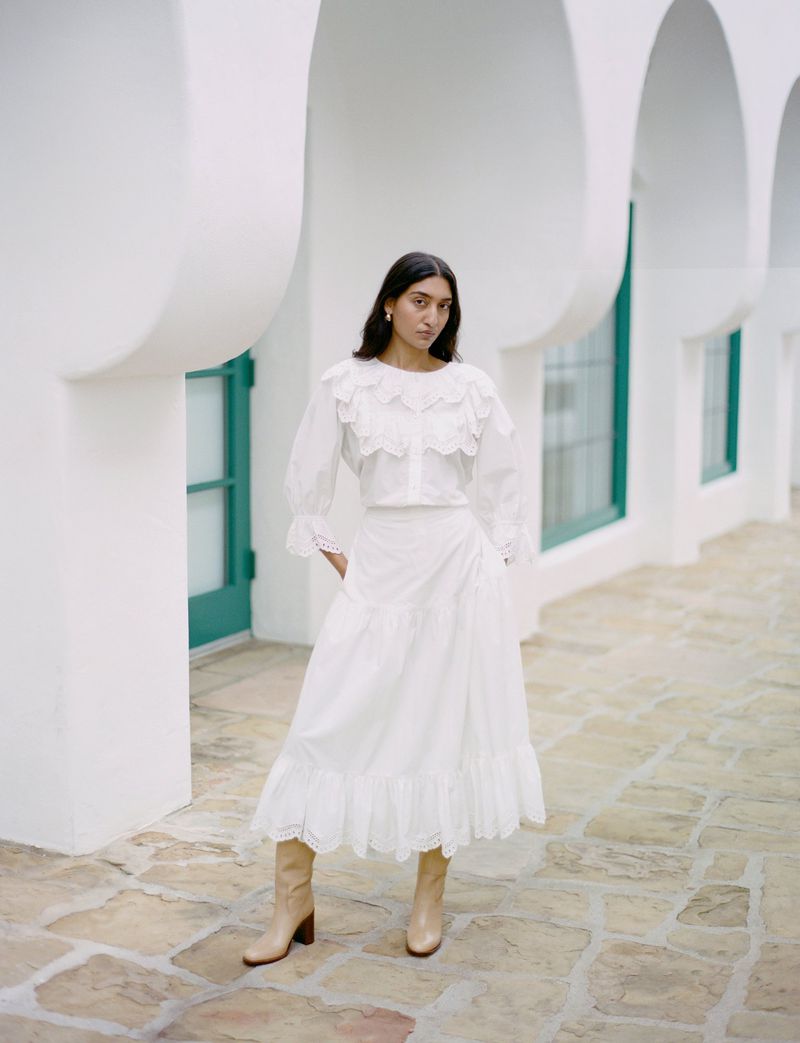 Woman poses in a white ruffled blouse and skirt, complemented by beige boots, against an outdoor setting with curved architectural details.