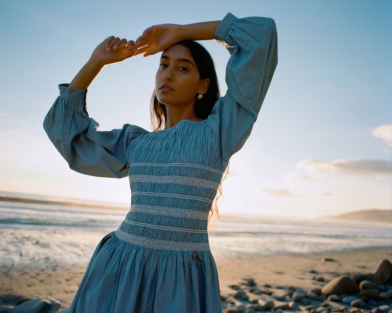 A south asian woman in a long-sleeved blue dress poses on a sandy beach with the ocean and a bright sky in the background.