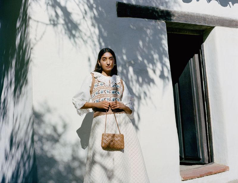 Woman in a white skirt and patterned sweater vest holding a woven handbag, standing against a white wall with tree shadow patterns.