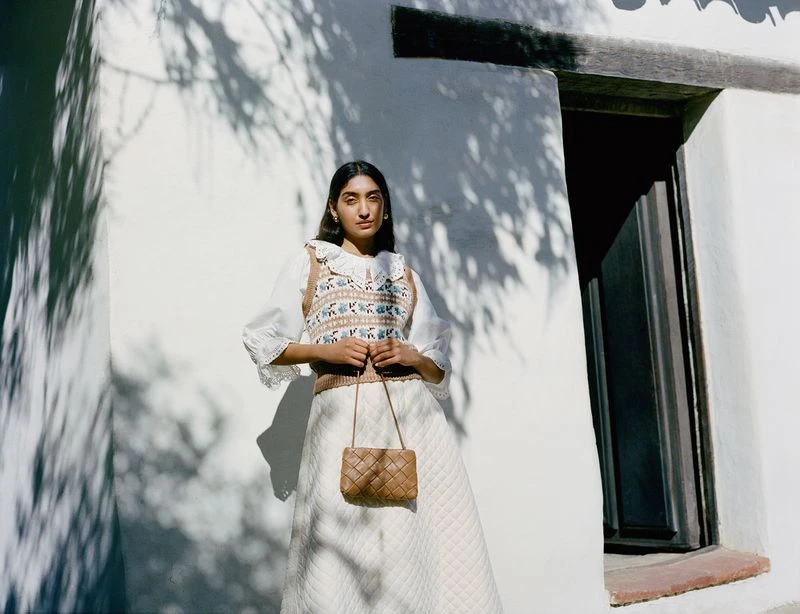 Woman in a white skirt and patterned sweater vest holding a woven handbag, standing against a white wall with tree shadow patterns.