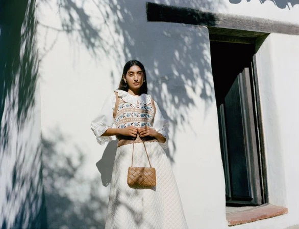 Woman in a white skirt and patterned sweater vest holding a woven handbag, standing against a white wall with tree shadow patterns.