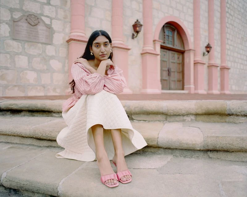An adult woman sits on stone steps wearing a pink blouse, white skirt, and pink sandals in front of a light-stone building with pink columns.
