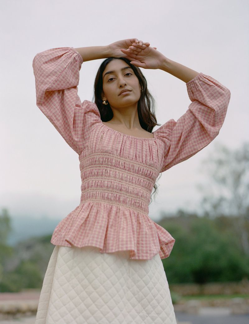 Brunette woman in pink gingham top and cream skirt poses outdoors with hands above head against a blurred natural background.