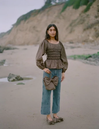 A woman in a brown smock top, jeans, and matching bow shoes and bag stands on a beach with a neutral-toned cliff in the background.
