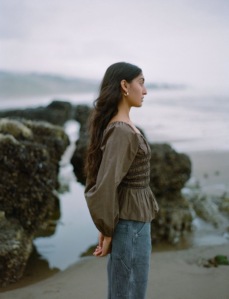 Woman in a brown gingham top and blue carpenter pants stands on a beach with large rocks and water in the background on an overcast day.