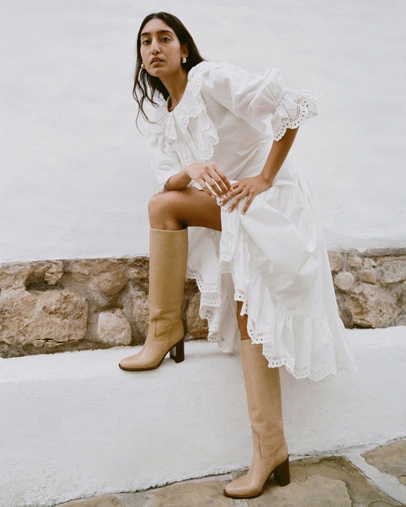 A stylish woman poses outdoors, wearing a white eyelet dress and tan knee-high boots, sitting on a stone ledge against a bright background.