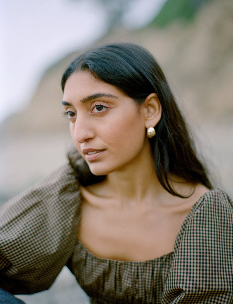 Close-up portrait of a dark-haired adult woman wearing a checkered top and gold earrings outdoors in natural light with a shallow depth of field.