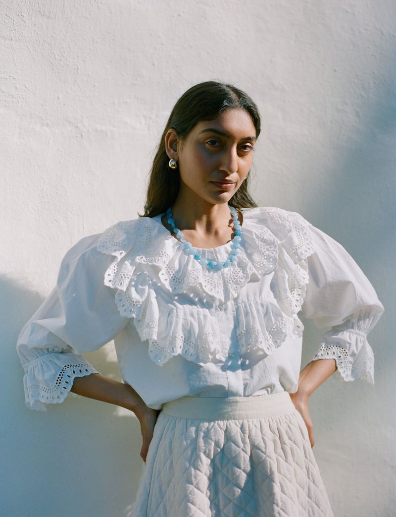 Model Lizzy Yusuff wearing white eyelet blouse and blue beaded necklace against textured wall
