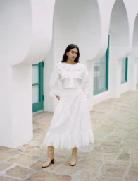 Model standing outdoors wearing a white ruffled blouse, white skirt, and tan boots under arched white porticos.