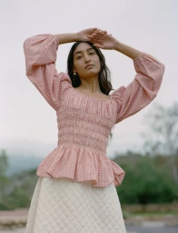 Model Lizzy Yusuff in pink gingham smocked top and cream skirt with arms raised against an outdoor backdrop, for Loeffler Randall FW24.
