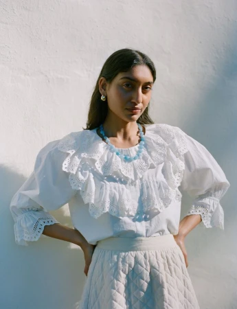 Model Lizzy Yusuff wearing white eyelet blouse and blue beaded necklace against textured wall