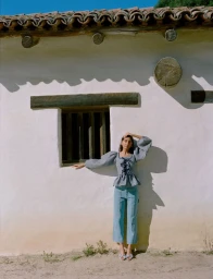 Model standing outdoors against a white stucco building exterior wearing a patterned puff-sleeve top and blue denim trousers.
