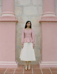 Model standing between two pink architectural columns wearing a pink patterned top and white quilted skirt.