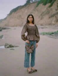 Model standing on a sandy beach wearing a brown smocked blouse, blue jeans, and holding a brown bow handbag for Loeffler Randall
