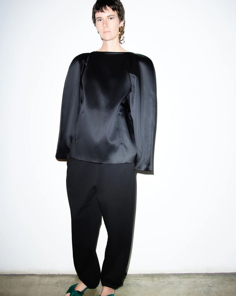 A woman in a black satin long-sleeve top and baggy black pants poses against a white backdrop, showcasing a minimalist fashion style.