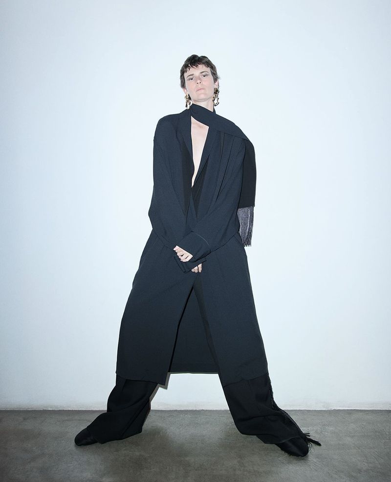 Model wearing a long dark coat and scarf against a plain wall for Carven SS25 Pre-collection