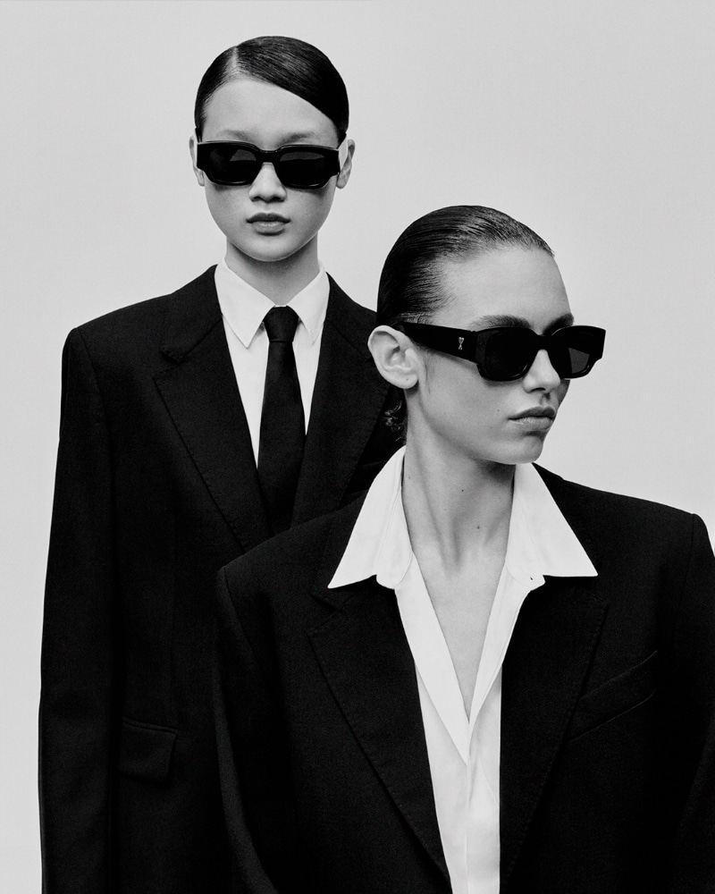Two models in black suits and sunglasses pose in a black and white studio shot, emphasizing fashion and minimalist style against a plain backdrop.