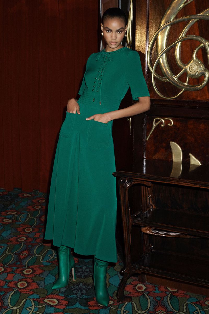 Elegant woman in a green dress and matching boots poses indoors next to a vintage wood cabinet with floral patterned flooring.