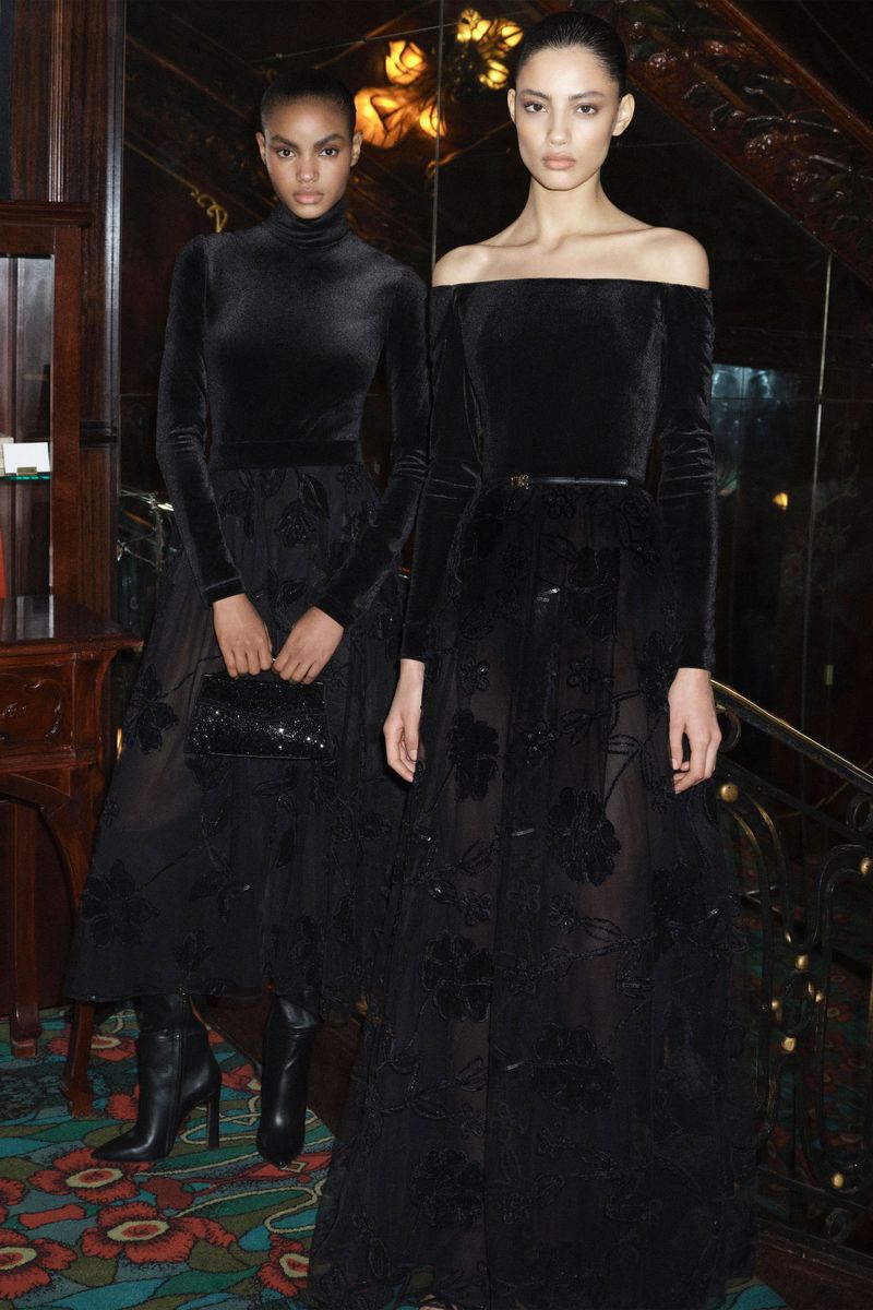 Two fashion models showcase elegant black gowns indoors, one with a turtleneck and the other off-the-shoulder, in a full shot.