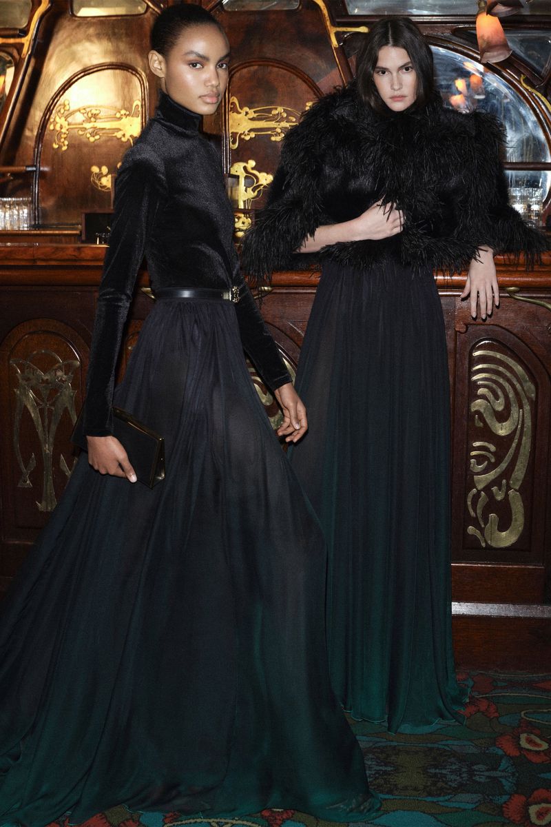 Two fashion models in elegant black gowns pose indoors against an ornate backdrop with dark, moody lighting and a touch of green.