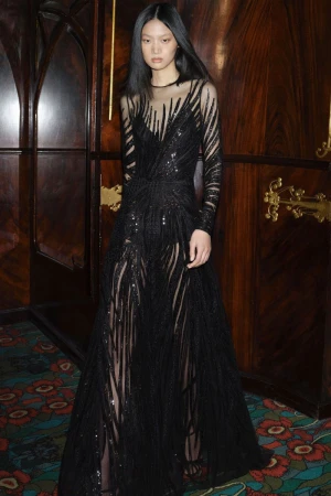 Model in a black embellished evening gown standing against dark wood paneling inside a room