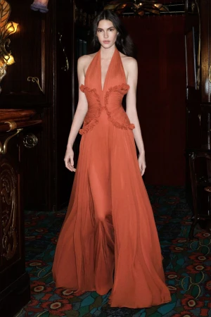 Model wearing a long rust-colored halter dress standing in a dimly lit interior with dark wood paneling and patterned carpet.