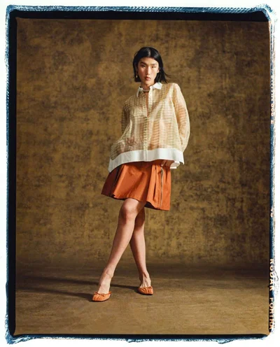 Model standing on a textured brown surface wearing a sheer patterned shirt and orange pleated skirt for 3.1 Phillip Lim.