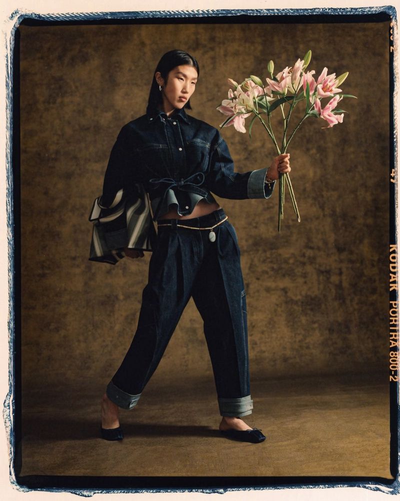A stylish Asian woman in denim attire poses indoors, holding pink lilies against a textured brown backdrop, in a full shot with soft lighting.
