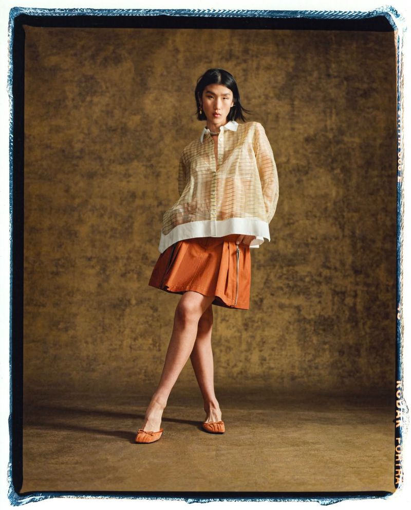 A stylish adult woman poses in a studio wearing an orange skirt and sheer blouse, standing against a textured brown backdrop.