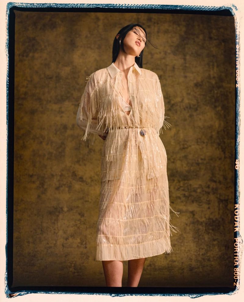 A stylish woman poses in a cream-colored fringed dress with dark hair, set against a textured gold background in a studio fashion shot.
