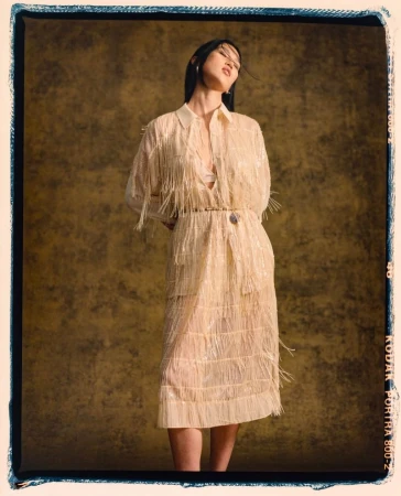 A stylish woman poses in a cream-colored fringed dress with dark hair, set against a textured gold background in a studio fashion shot.