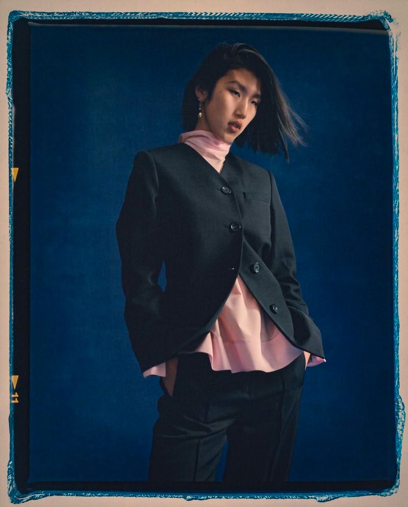 Portrait of a fashionable young adult woman in a black suit and light pink top against a dark blue backdrop in a studio.